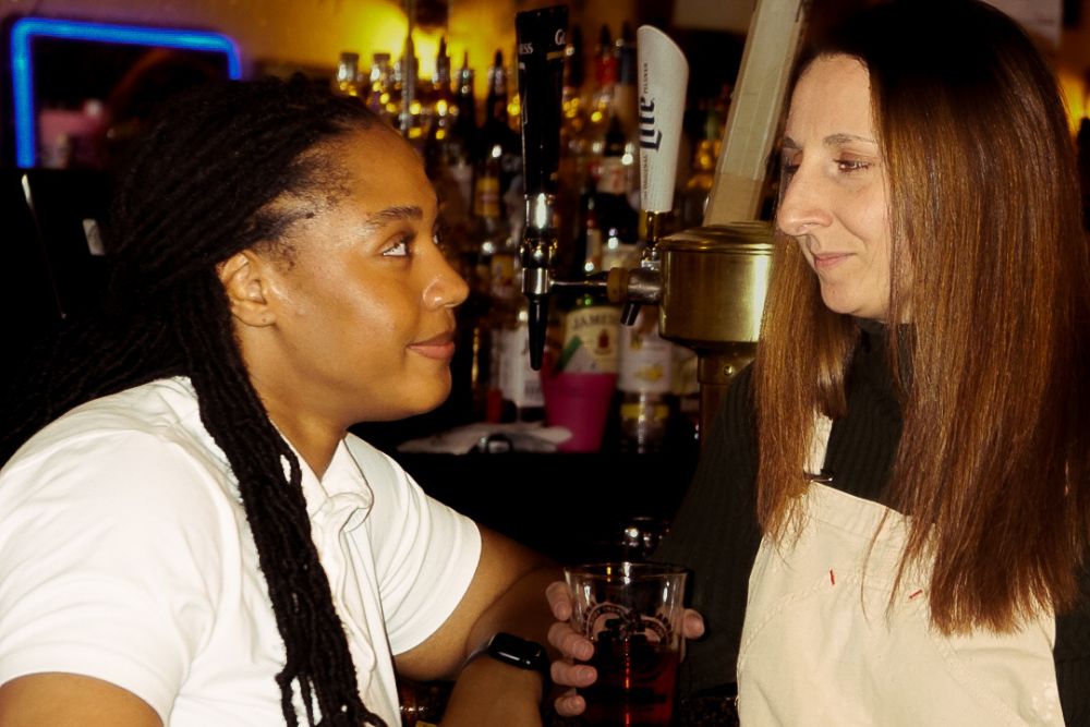 Two women smile at each other in a warmly lit bar. The woman on the right holds a drink.