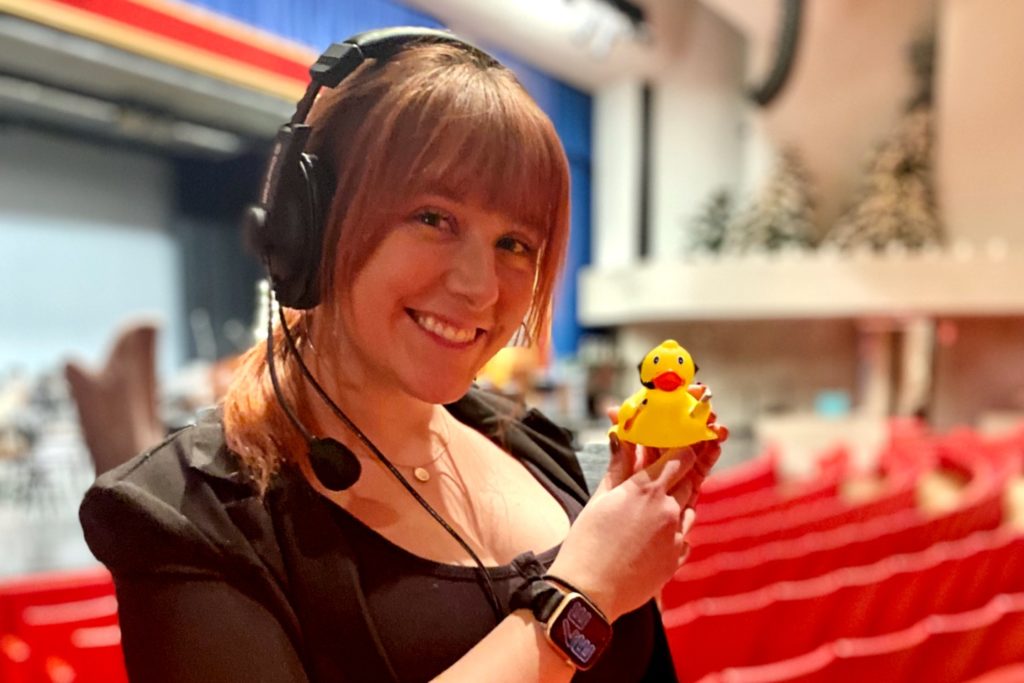 Woman wearing a headset with microphone holding a rubber duck that also has a headset and microphone on it.