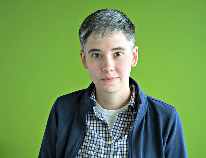 A picture of a person wearing a black coat and checkered shirt in front of a green background. 
