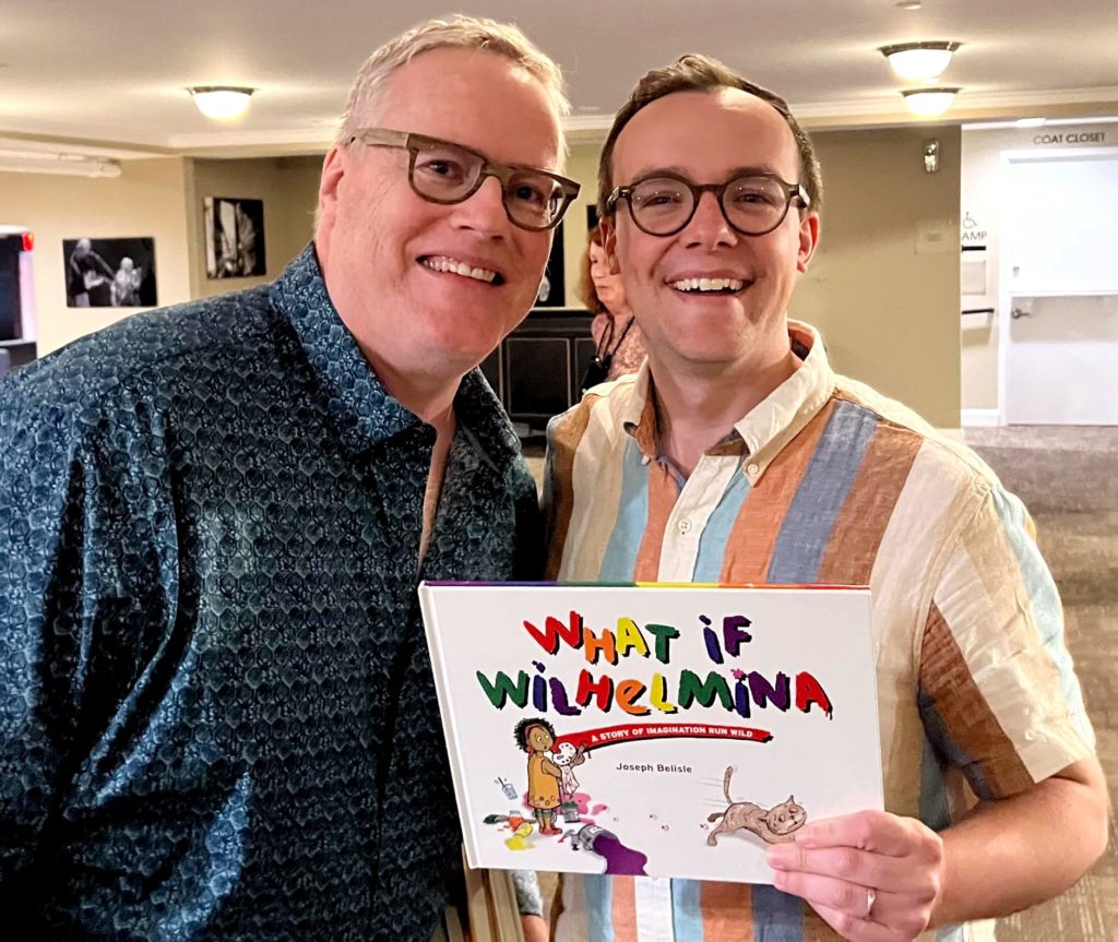 Photo of two men standing next to each other and hold a book with the title "What if Wilhelmina" on the cover. Both men are white males and both are wearing glasses and are smiling. 