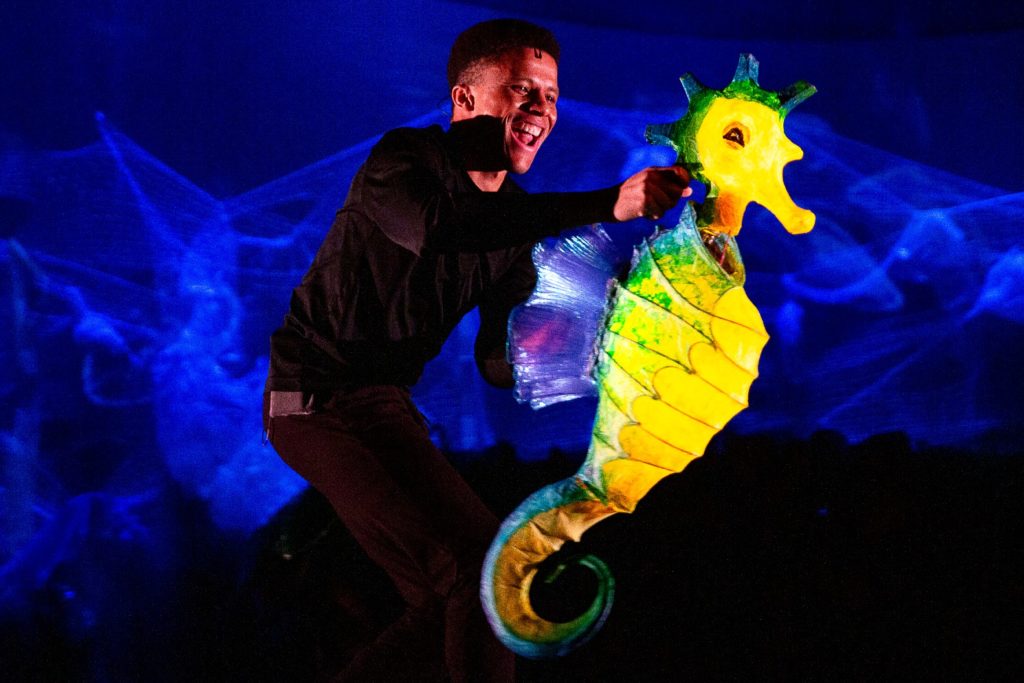 Evan Hill Philips on stage holding a large seahorse puppet. 
