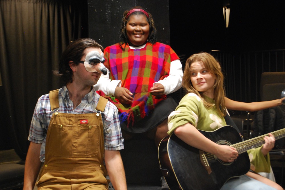 Three people - one wearing a face mask, one playing a guitar, one standing and smiling. 
