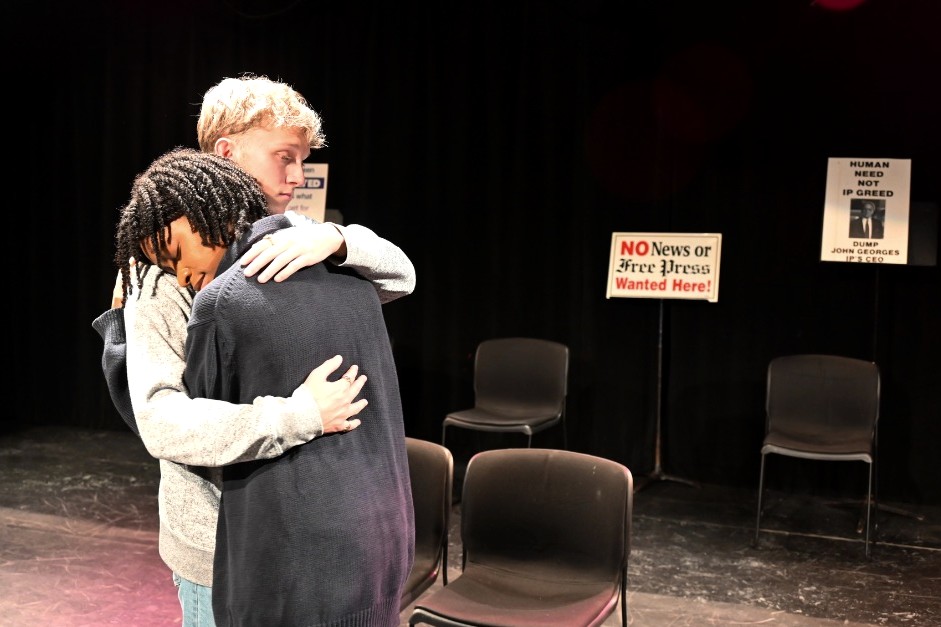 Two actors, a boy and a girl, hugging on stage with a sign in the background that says: "No News of Free Press Wanted Here!"