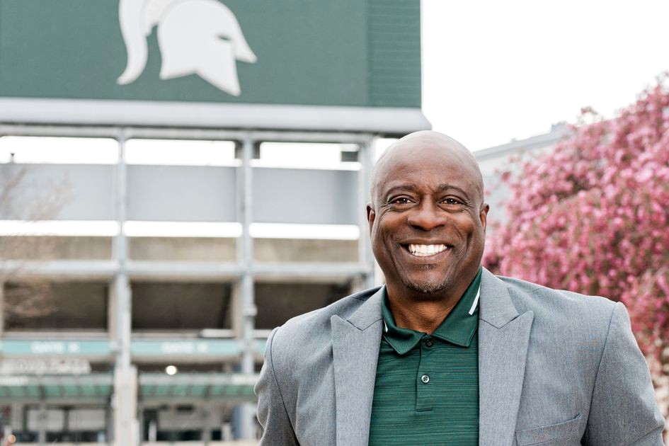 A smiling man in a green polo and gray blazer stands in front of Spartan Stadium at Michigan State University, with the Spartan helmet logo visible behind him and pink flowering trees in the background.